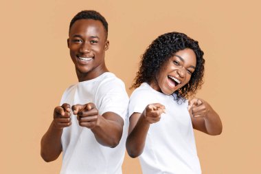 Neşeli genç Afrikalı Amerikalı erkek ve kadın kamerayı işaret ediyor, sarı stüdyo arka planı, boş alan. Oynayan genç siyah çift flört ediyor, kameraya parmak tabancalarını gösteriyorlar.