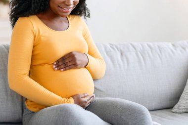 Mutlu Afro-Amerikalı kadın çocuk bekliyor, evde vakit geçiriyor, boş bir alan. Oturma odasındaki koltukta oturan neşeli zenci hamile kadın, sarılıyor ve koca göbeğine bakıyor.