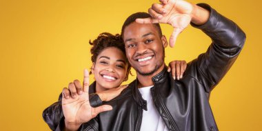 Yüzlerini çerçeveleten, sarı stüdyo arkaplanının üzerinde siyah deri ceketler giyen neşeli Afrikalı Amerikalı çift. Panorama