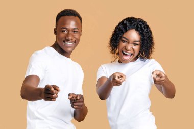 Oynaşan genç siyahi çift kameraya parmak tabancalarını gösteriyor. Neşeli genç Afrikalı Amerikalı erkek ve kadın arka arkaya kamerayı işaret ediyor, sarı stüdyo arka planı, boş alan