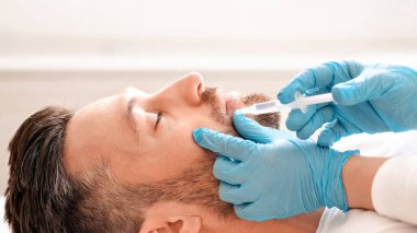 Mavi eldivenli güzel eller yakışıklı sakallı adam için ağız dolgusu yapıyor. Çekici orta yaşlı iş adamı estetik kliniğinde burun güzelliği iğnesi yaptırıyor.
