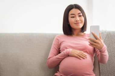 Hamile Çinli Kadın 'ın telefonuyla evdeki koltukta selfie çekerken mutlu musun? Hamilelik Tatil Konsepti. Asyalı Anne Hamilelik sırasındaki akıllı telefonuyla kendi portresini yapıyor.