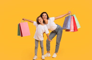 Alışveriş ve Satış. Neşeli Arap Anne ve Kızı Sarı Arkaplanda Renkli Alışveriş Çantaları Tutuyor. Orta Doğu Aile Reklamları harika bir teklif. Stüdyo Çekimi