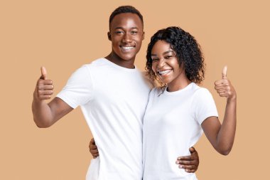 Sarı stüdyo arka planında sarılıp baş parmağını gösteren iki mutlu zenci aşık, panorama. Neşeli Afro-Amerikalı kadın ve erkek kucaklaşıp el kol hareketi yapıyorlar, ürün, hizmet veya alım öneriyorlar.