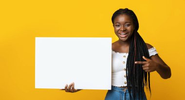 Beyaz boş levhaya işaret eden neşeli siyahi kadın, mesajınız ya da ilanınız için bedava yer gösteriyor, reklam panosunda kopya alanı gösteriyor, sarı stüdyonun arka planında poz veriyor.