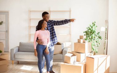 Afrikalı Amerikalı adam mutlu kız arkadaşına bir şey gösteriyor, yeni evlerinde kutuların arasında duruyor, boş alan. Pozitif siyah bir adam taşınma gününde karısına sarılıyor.