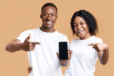Genç, duygusal, Afro-Amerikalı çift boş bir siyah akıllı telefon ekranını işaret ediyor, sarı stüdyo geçmişi var. Gülümseyen siyah adam ve kadın cep telefonlarında güzel bir uygulama gösteriyorlar.