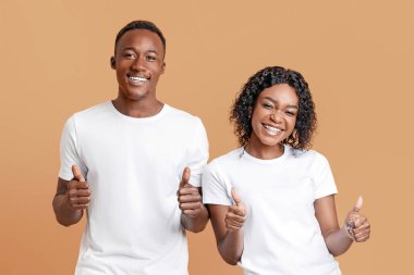 Emotional black lovers showing their thumbs up on yellow studio background. Cheerful young african american man and woman recommending product, service or purchase, giving likes