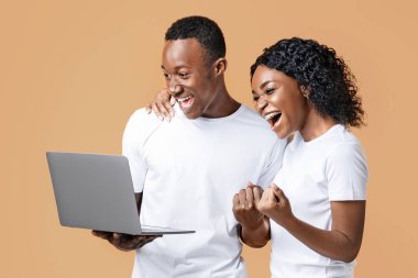 Positive african american millennial man and woman celebrating win, holding laptop, shouting and gesturing on yellow studio background. Young black couple gambling online successfully, using laptop