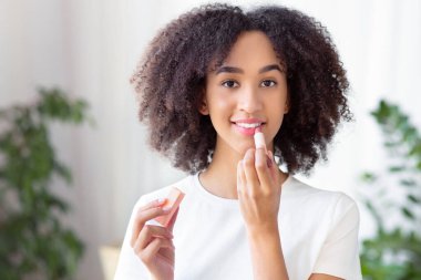 Evde dudaklarla ve güzellikle ilgilenmek. Kıvırcık saçlı mutlu Afro-Amerikan kadın nemlendirici ruj sürüyor ve sabahları oturma odasında bitkilerle kameraya bakıyor.