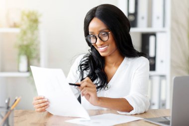 Ofisteki masasında belgeye bakan çekici siyahi iş kadını. Afrika kökenli Amerikalı kadın sekreter evrak işleri, okuma sözleşmesi veya iş yerinde sözleşme yapıyor.