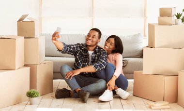 Gülümseyen siyah adam ve kız arkadaşı kendi evlerine taşınıyorlar, karton kutuların arasında selfie çekiyorlar, panorama. Mutlu Afrika kökenli Amerikalı çift yeni dairedeki ilk günün fotoğrafını çekiyor.