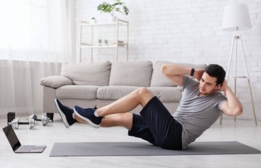 Spor ve sağlıklı yaşam tarzı. Adam ellerini başının arkasına koyup karın kasları için egzersiz yapıyor ve evde iç mekanda online eğitim izliyor.