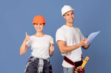 Tamir Servisi Ekibi. Mavi arka planda, Hardhat şapkalı ve boya fırçalı mutlu adam ve kadın, gülümseyen kadın başparmağını kaldırıyor, evi yenilemeye hazır, kopyalama alanı var.
