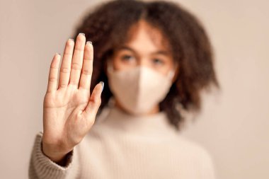 Grip ve bulaşıcı salgın sırasında sağlığın korunması ve önlenmesi veya virüs ve salgın hastalıkların durdurulması. Bulanık maskeli Afrikalı Amerikalı kadın açık arkaplanda CV19 'a el hareketi yapıyor.