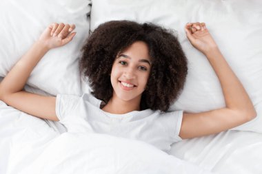 Hafta sonu, tatil ve evde dinlenme. Coperid-19 sırasında. Mutlu Afrikalı genç Amerikalı bayan yatağa uzanıyor, kameraya bakıyor ve rahat bir yatak odasında dinleniyor.