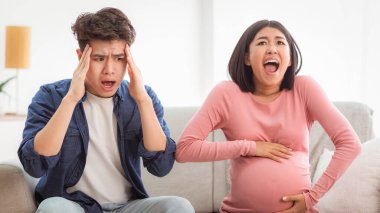 Hamilelik sorunları. Hamile Asyalı Eş Karına Spazm Geçirirken Bağırıyor, Koca Evde Koltukta Otururken Başa Dokunuyor. Çocuk Doğumu Konusu, Ani Çalışma Konsepti