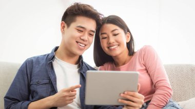 Genç Asyalı koca ve karısı evdeki oturma odasındaki koltukta otururken internette dijital tablet kullanarak internete bakıyorlardı. Hafta sonu tatili, iyi seyirler dilerim. Modern Gadget Yaşam Biçimi