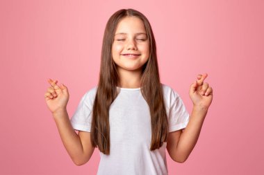 Sürpriz bekliyoruz. Gözleri kapalı ve parmakları çapraz gülümseyen çocuk dilek tutar, pembe arka planda izole edilmiş, stüdyo çekimlerinde.