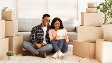 Tablet bilgisayarlı neşeli siyah çift taşınma gününde karton kutuların arasında oturuyor. Yeni ev sahipleri evleri için ev eşyaları satın almak için internette geziniyorlar. Panorama