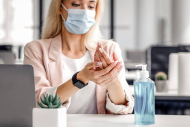 Koronavirüs salgını sırasında yeni normal ve hijyen. Akıllı saatli koruyucu maskeli kız sağlıkla ilgilenir, iş yerindeki elleri dizüstü bilgisayarla dezenfekte eder, boş alan yaratır.