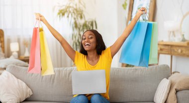 Dizüstü bilgisayarı olan heyecanlı zenci kadın evdeki kanepede alışveriş torbalarını tutuyor. Güzel Afrikalı Amerikalı kadın oturma odasında internetten mal alıyor.