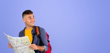 Sırt çantası haritalı neşeli bir erkek turist mavi stüdyo arka planındaki boş alana bakıyor. Yakışıklı gezgin tatilini ya da yolculuğunu planlıyor, rotasını seçiyor.