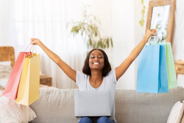 Evdeki kanepede elleri alışveriş torbalarıyla dolu dizüstü bilgisayarı olan heyecanlı siyahi kadın. Sevimli Afrikalı Amerikalı kadın internet dükkanında büyük indirimler yaptığı için mutlu.