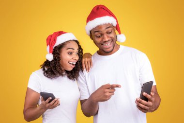 Noel Mobil Uygulaması. Akıllı Telefon Uygulaması Öneren, Sarı Stüdyo Arkaplanı Üzerine Noel Baba Şapkası Giyen Heyecanlı Siyah Çift Cep Telefonu Tutuyor. Xmas Reklamcılık. Panorama