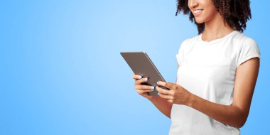 Modern cihazlarla çalışmak ve rahatlamak. Kıvırcık saçlı, internette sörf yapan, pembe tişörtlü, gülümseyen Afro-Amerikalı kadın menekşe arka planında izole edilmiş dijital tablet kullanıyor.