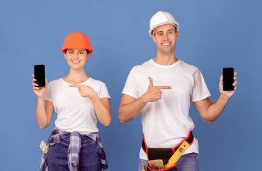 Koruyucu miğferli mutlu çift siyah ekranlı akıllı telefonları işaret ediyor, ev yenileme uygulaması veya inşaat malzemeleri web sitesini öneriyor, mavi stüdyo arka planının başında duruyor,