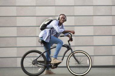 Sırt çantalı ve kulaklıklı neşeli siyah adam bisiklet sürüyor. Tuğla duvarın yanında.