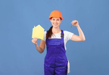 Kapaklı ve Hardhat 'lı Mutlu Kadın Anahtarları ve Sarı Kağıt Ev Figürü, Güvenilir Bina ve Onarım Hizmetleri Öneren Olumlu Kadın Mavi Stüdyo Arkaplanının Üzerinde Duran, Boşluğu Kopyalayan