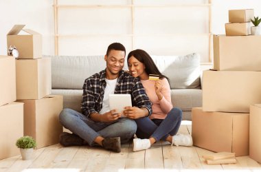 Genç siyah çift taşınma günü yeni evlerinde online alışveriş yapmak için dizüstü bilgisayar ve kredi kartı kullanıyor. Kız arkadaşıyla birlikte internetten ev eşyaları alan Afro-Amerikalı bir adam.