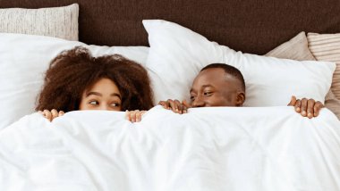 Rahat yatakta romantizm ve eğlence, hafta sonları evde mutlu bir çift. Neşeli Afro-Amerikan kadın ve erkek milenyumu beyaz battaniyenin altına bakıyorlar ve birbirlerine bakıyorlar, boş alan, üst manzara.