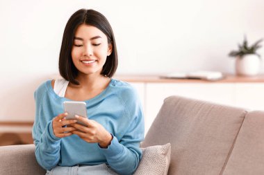 Evde vakit geçirmek. Gülümseyen Asyalı genç kadın cep telefonunu kullanıyor, internete bakıyor, sosyal medyayı kontrol ediyor oturma odasındaki kanepede oturuyor. Mutlu kadın arkadaşlarına mesaj atıyor, internetten video izliyor.