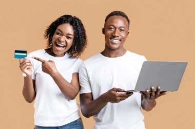 Çevrimiçi bankacılık ve e-ticaret. Mutlu Afro-Amerikalı erkek ve kadın kredi kartını gösteriyor ve sarı stüdyo geçmişi üzerinde çalışıyor. Siyah çift online alışveriş yapıyor, dizüstü bilgisayar kullanarak.