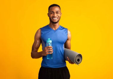 Fitness Konsepti. Elinde su şişesi ve yoga minderi tutan mutlu siyah adam, sarı duvarın üzerindeki kameraya gülümsüyor.