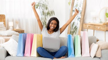 İnternetten alışveriş yapan heyecanlı siyahi kadın, dizüstü bilgisayarlı koltukta oturuyor, hediye çantalarıyla çevrili, kapalı bir yerde. Güzel Afrikalı Amerikalı kadın internette hediyeler alıyor, büyük indirimler alıyor.