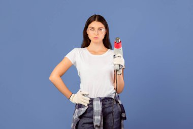 Yenilemeye hazırım. Koruyucu gözlük takan, inşaat kurutma makinesini tutan, mavi stüdyonun arka planında duran, ciddi tamirci ve tamirci bir çift portresi.