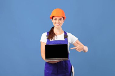 Örtüdeki Gülümseyen Bayan ve Sert Şapkalı Siyah Ekran ile dizüstü bilgisayara işaret eden, Çevrimiçi alışveriş için yeni web sitesi öneren, mavi arka planda dururken internetten inşaat araçları alan,