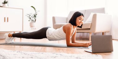 Evde antrenman. Laptop bilgisayarında online özel ders seyrederken tahtayla oynayan, yoga minderinin üzerinde oturma odasında egzersiz yapan Asyalı bir kadının tam boy portresi.