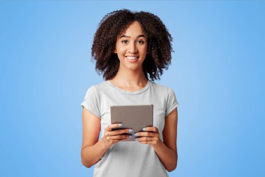 İnternette sörf yapmak için boş zaman ve cihazlar. Bin yıllık neşeli Afro-Amerikan kadın pembe tişörtlü, kıvırcık saçlı, tablet tutan, internetten alışveriş yapan ve kameraya bakan, boş alan