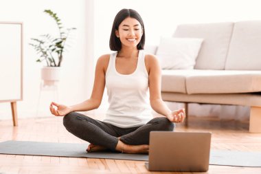 Sanal Meditasyon. Lotus pozisyonunda oturmuş yoga ve nefes egzersizi yapan Asyalı sakin bir bayan. Sağlık sigortası kavramı. Oturma odasında dizüstü bilgisayarda ders izleyen bir kadın.
