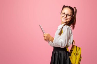 Çocuk çalışmak için aletler kullanıyor. Sırt çantalı neşeli liseli kız dijital tablet tutuyor ve pembe arka planda kameraya bakıyor.