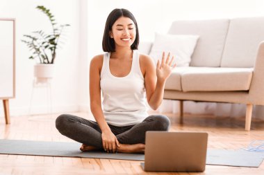 Çevrimiçi ev eğitimi. Spor Asyalı bir kadının webcam 'e el salladığı, yoga minderinin üzerinde dizüstü bilgisayarın önünde oturduğu portresi. Bilgisayarda ders izleyen ya da kayıt yapan bayan, oturma odasında egzersiz yapıyor.