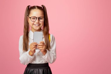 Modern teknoloji ve okul. Gözlüklü gülümseyen küçük kız elinde akıllı telefon, pembe arka planda izole edilmiş, stüdyo çekimi, boş alan