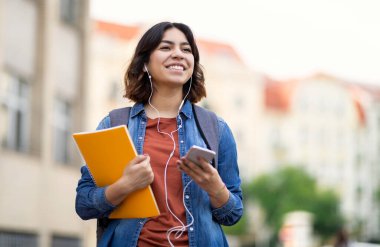 Neşeli genç Arap kız öğrenci dışarıda akıllı telefon ve kulaklıkla müzik dinliyor, çalışma kitapları ve sırt çantası taşıyan orta doğulu kadın şehir sokaklarında yürürken gülümsüyor, boşluğu kopyalıyor.