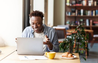 Kahve dükkanındaki dizüstü bilgisayardan iletişim kuran kulaklıklı mutlu siyah adam.