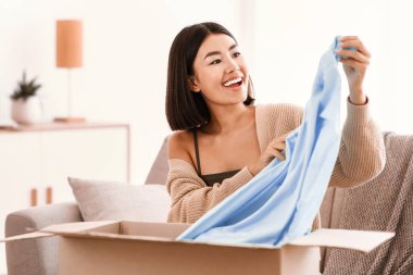 Posta Teslimatı, Sevkiyat ve İnsanlar Konsepti. Mutlu Asyalı kadın paketleri açıyor, kanepeye oturuyor ve elbiselerini tutuyor, karton paketleri açıyor, fotokopi çekiyor. Satın almaktan memnun bayan